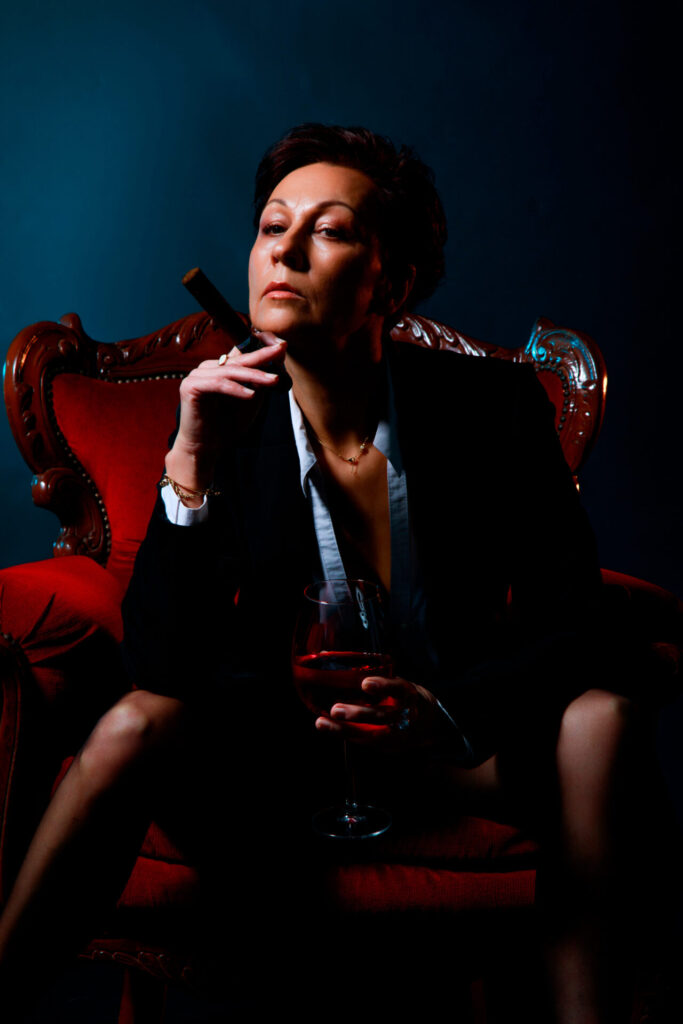 Portrait boudoir en smoking : femme assise dans un fauteuil rouge, verre de vin à la main, éclairage contrasté